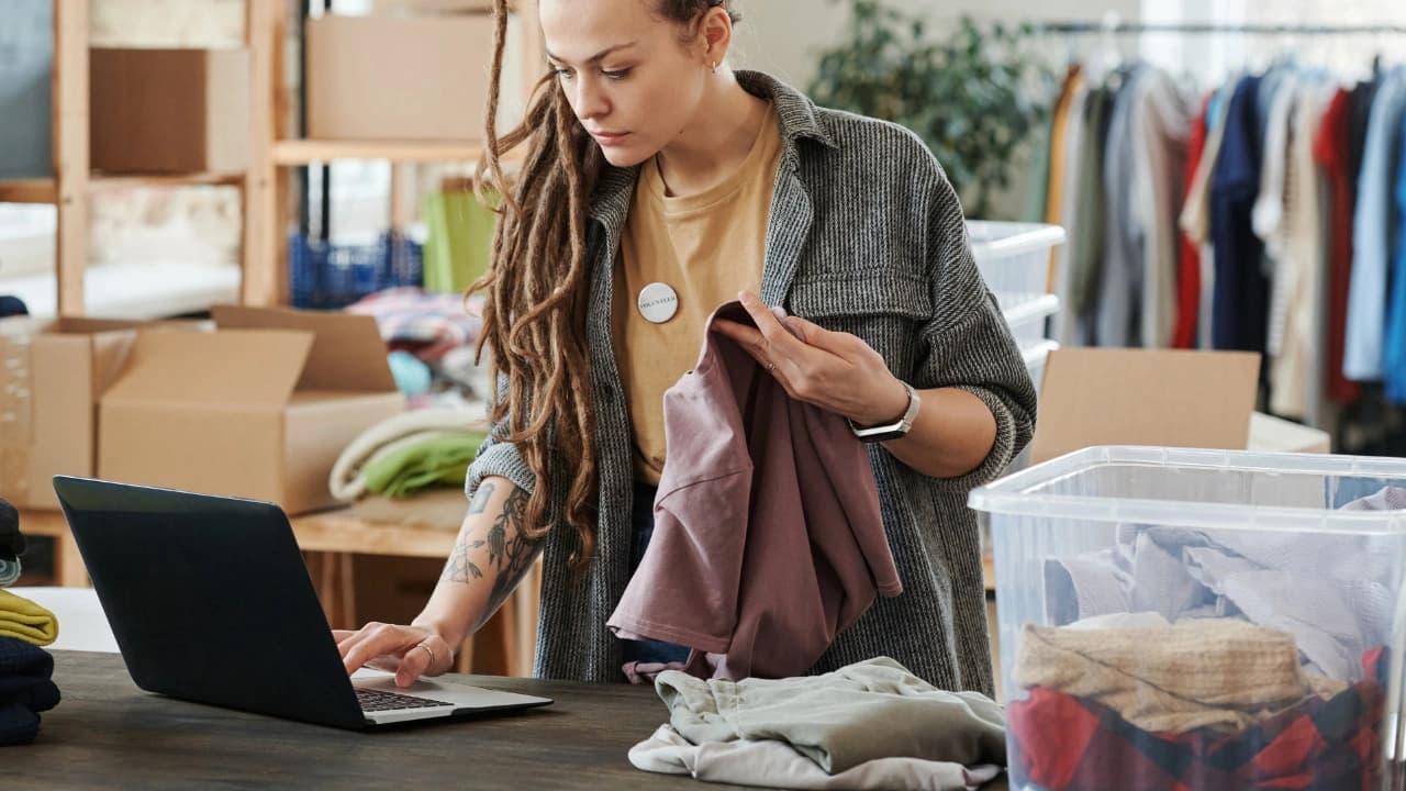 Woman with dreadlocks looking at laptop, holding fabric, managing custom t-shirt design and apparel orders.