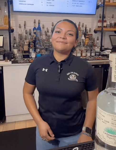 Bartender in a black branded polo shirt behind a liquor display, illustrating staff apparel marketing and the impact of custom restaurant staff uniforms in turning uniforms into wearable brand assets.