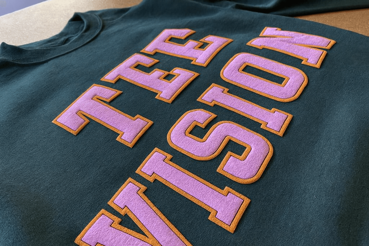 This dark green sweatshirt features bold, block lettering that spells out “TEE VISION” in pink with a brown outline, showcasing the dimensional effect that defines puff print apparel. For anyone asking what is puff printing, this image serves as a visual example of puff ink screen printing in action, where the ink expands to create raised ink printing that you can both see and feel. The textured ink printing highlights how custom puff printing transforms a simple sweatshirt into a premium piece, often used in streetwear puff designs to add depth and tactile appeal. Compared to flat screen printing techniques, puff vs flat printing demonstrates how much more dynamic and eye-catching puff can be, making it a staple in modern screen printing techniques for fashion-forward brands.