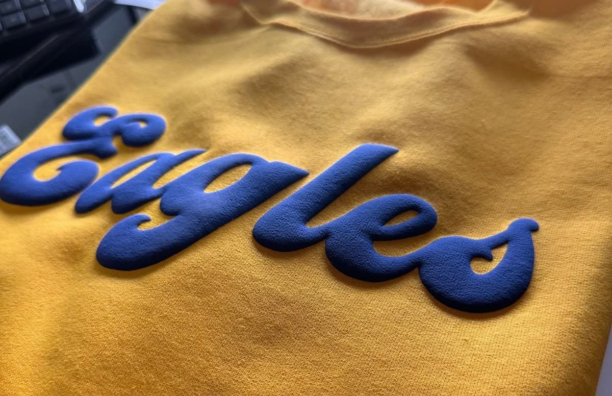 The yellow sweatshirt with raised blue “Eagles” script is a clear example of what is puff printing, where puff ink screen printing produces textured ink printing that stands out. This puff print apparel shows how custom puff printing enhances team branding with bold, dimensional lettering.