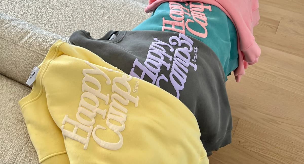 Three “Happy Camper” sweatshirts in different colors demonstrate branding consistency while connecting to what is puff printing. Although embroidered, the raised effect mirrors puff print apparel, showing how textured ink printing and puff vs flat printing both add dimension to lifestyle wear.