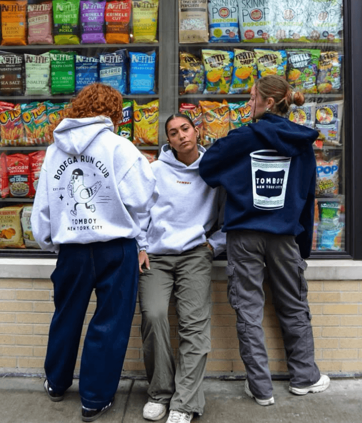 Tomboy Athletic Club hoodies with coffee cup graphics photographed outside New York City bodega, blending streetwear style with urban culture.