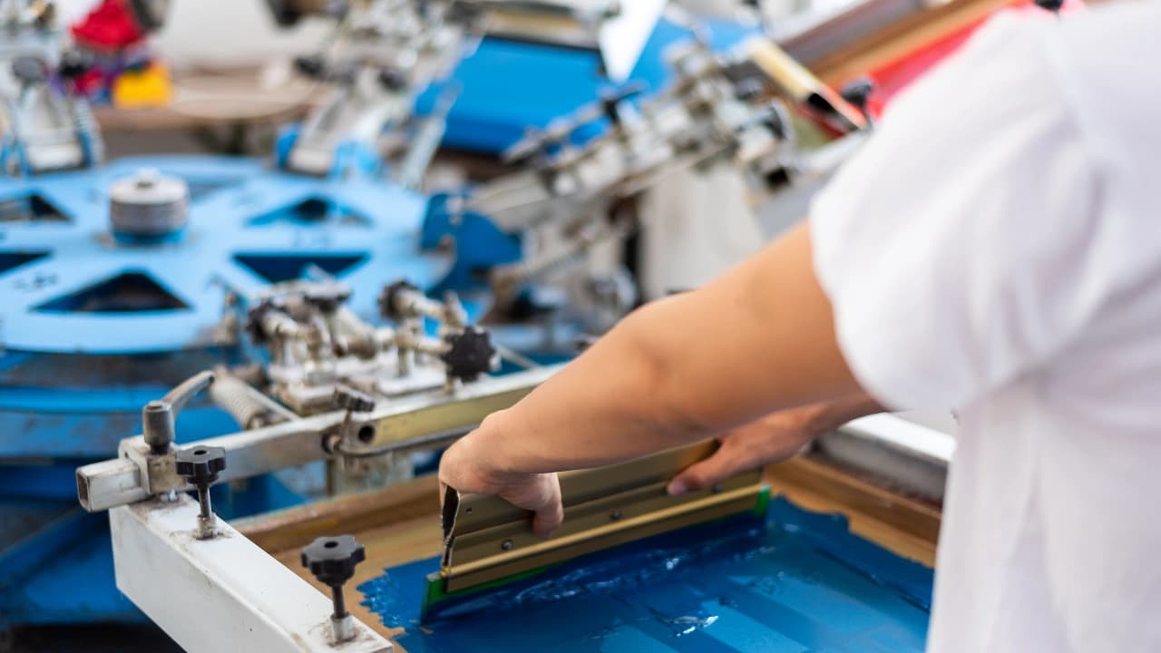A worker performs custom t-shirt silk screen printing, pulling blue ink across a screen for an order of bulk shirts.