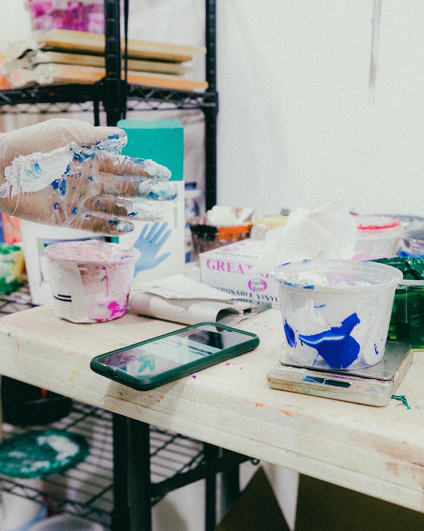 Hands in messy gloves, surrounded by paint and a phone, showing custom t-shirt design and screen printing.